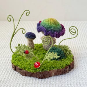 May include: A whimsical crochet fairy garden scene. It features a large mushroom with a rainbow-colored cap and purple ruffled edges, a smaller blue mushroom, and green foliage on a mossy base. A red ladybug and mushroom add pops of color.