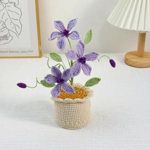 以下が含まれることがあります： 手作りのかぎ針編みのフラワーアレンジメント。紫色の花3つと緑の葉とつぼみがあり、黄色の縁が付いたベージュのかぎ針編みのポットに入っています。花は詳細で質感のある外観で、ポットはスカラップエッジです。