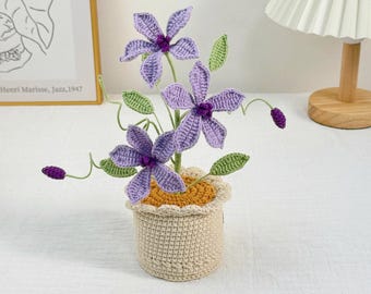 May include: A handmade crochet flower arrangement featuring three purple flowers with green leaves and buds, potted in a beige crocheted pot with a yellow rim. The flowers have a detailed, textured appearance, and the pot has a scalloped edge.