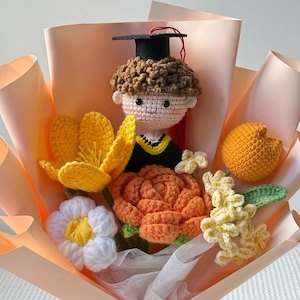 May include: A crocheted graduation bouquet featuring a doll wearing a graduation cap and gown, surrounded by crocheted flowers in yellow, orange, and white. The bouquet is wrapped in peach-colored paper.