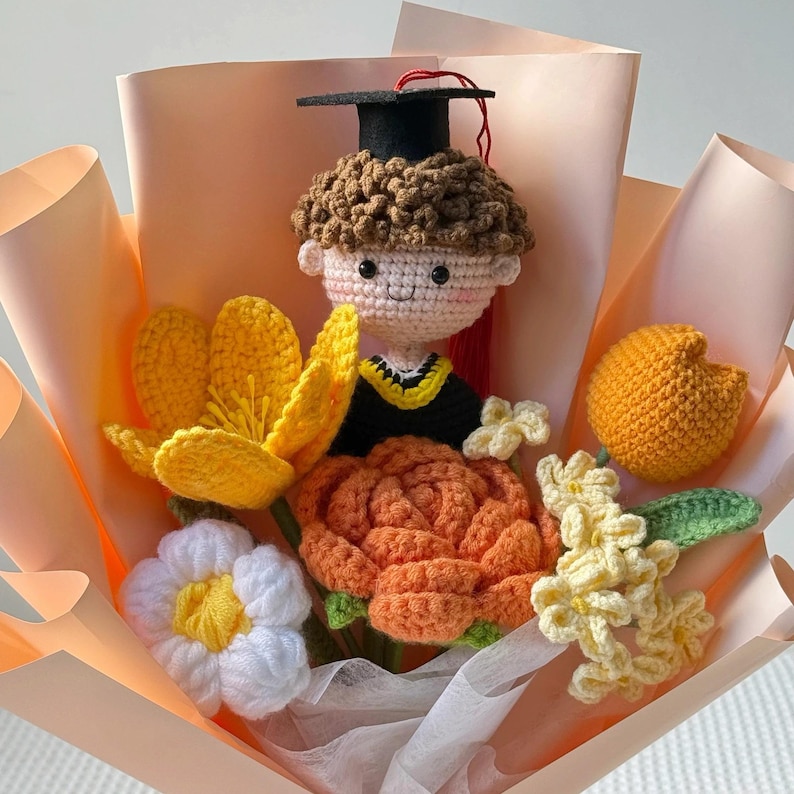 May include: A crocheted graduation bouquet featuring a doll wearing a graduation cap and gown, surrounded by crocheted flowers in yellow, orange, and white. The bouquet is wrapped in peach-colored paper.