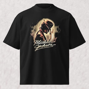 May include: A black short-sleeved t-shirt featuring a graphic print of Michael Jackson in a fedora and a sequined glove. The design includes a glowing light effect around the figure and the name Michael Jackson written in a cursive script font.