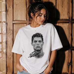 May include: A white short-sleeved t-shirt featuring a black and white photographic portrait of a man with styled hair on the chest area, accompanied by a bold black signature graphic positioned directly below the portrait on the front of the shirt.