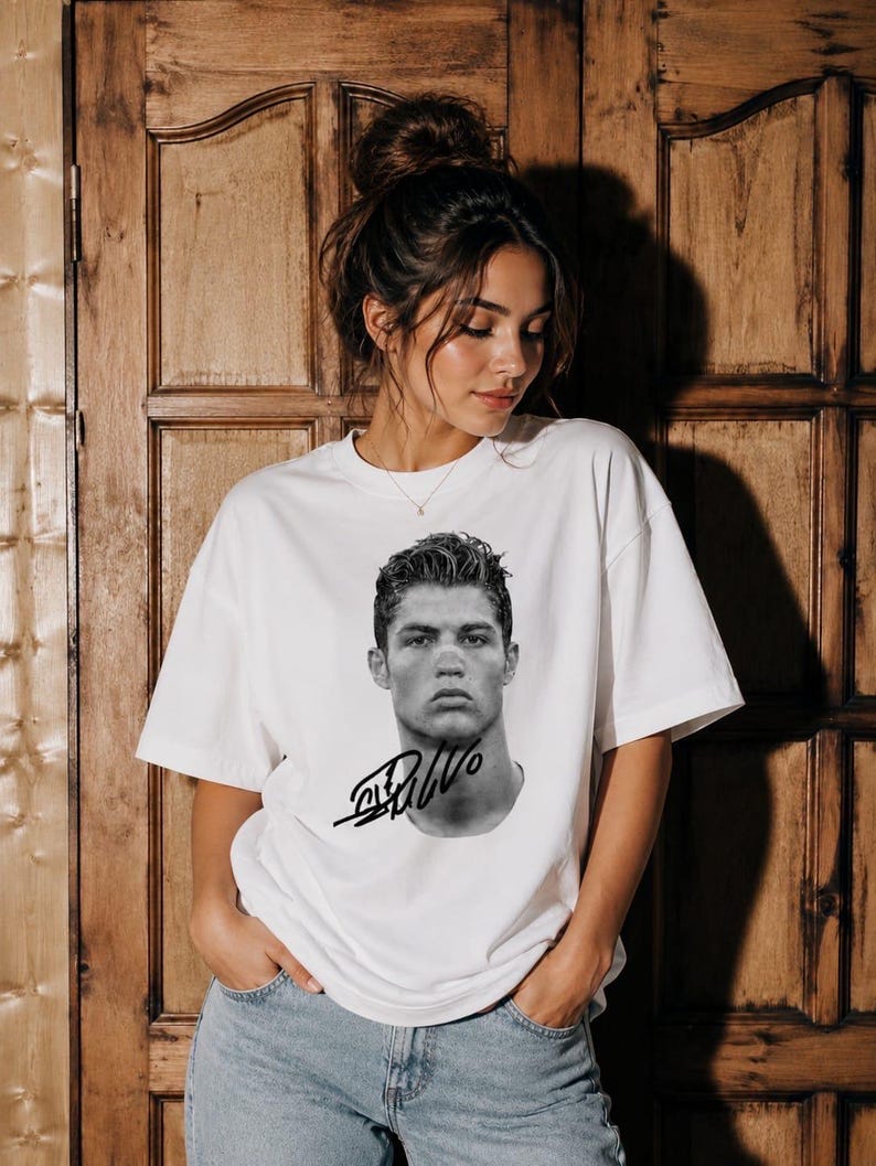 May include: A white short-sleeved t-shirt featuring a black and white photographic portrait of a man with styled hair on the chest area, accompanied by a bold black signature graphic positioned directly below the portrait on the front of the shirt.