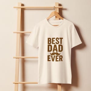 Puede incluir: Camiseta color crema con el texto marrón "BEST DAD EVER" y un bigote marrón. La camiseta está colgada en una percha de madera. El fondo es claro, lo que sugiere un estilo informal.