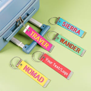 May include: A collection of colorful luggage tags with text, including "SIERRA", "WANDER", "NOMAD", and "TRAVER". Each tag is a different color, with a metal ring for attachment. One tag reads "Your Text/Logo".