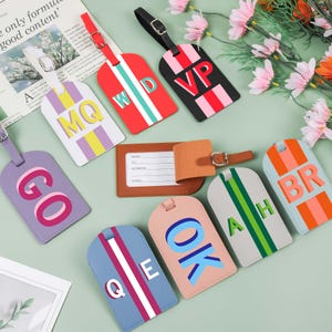 Könnte beinhalten: Eine Sammlung farbenfroher Gepäckanhänger mit verschiedenen Buchstabenkombinationen. Die Anhänger sind in Rot-, Schwarz-, Blau-, Rosa- und Lilatönen gehalten, mit weißen, grünen und orangenen Akzenten. Jeder Anhänger hat einen Riemen.