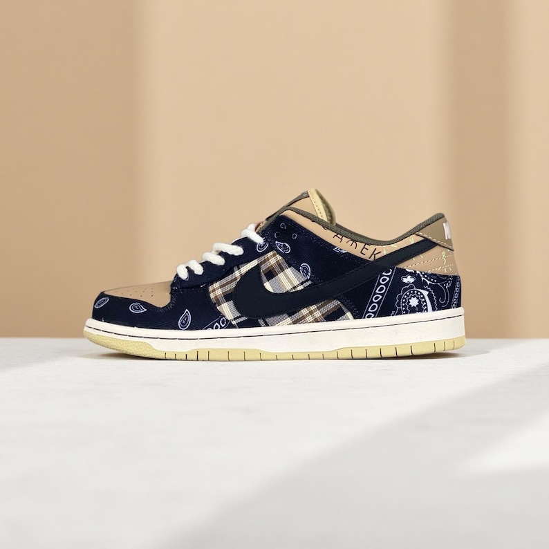 K&ouml;nnte beinhalten: Ein Nike Dunk Low Sneaker mit einer beigefarbenen Zehenpartie, marineblauen Seitenteilen mit Paisley-Muster und einem schwarzen Nike Swoosh. Die Ferse hat ein beigefarbenes Panel mit dem Aufdruck "AMEK". Die Sohle ist cremefarben.