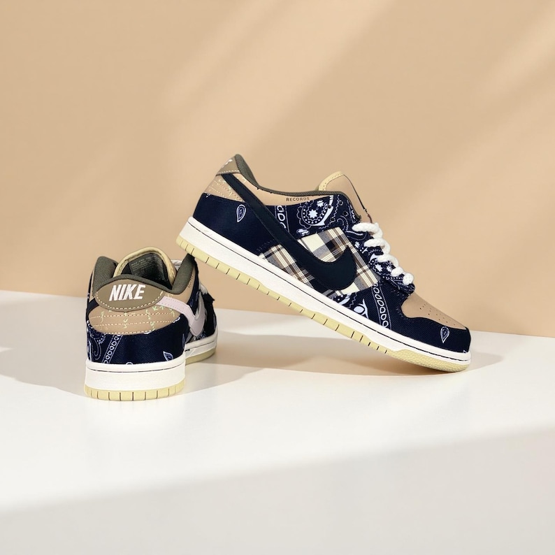 K&ouml;nnte beinhalten: Ein Paar Nike Dunk Low Sneaker. Die Schuhe haben eine marineblaue und beige Farbgebung mit einem Paisley- und Karomuster. Der Nike Swoosh ist schwarz. Die Sohlen sind cremefarben.