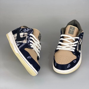 K&ouml;nnte beinhalten: Paar beige und marineblaue Sneaker mit wei&szlig;en Schn&uuml;rsenkeln. Die Sneaker haben ein Paisley-Muster auf den marineblauen Abschnitten und ein beiges Wildleder-&auml;hnliches Material auf der Spitze und der Zunge. Die Sohlen sind cremefarben.