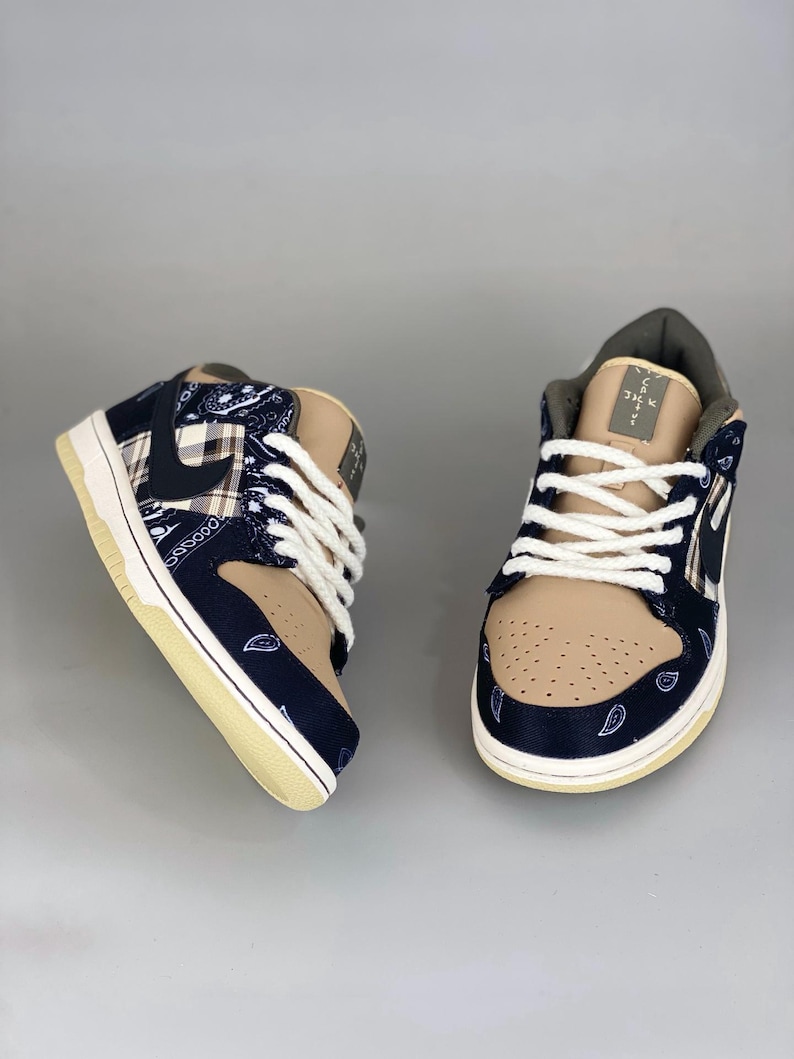 K&ouml;nnte beinhalten: Paar beige und marineblaue Sneaker mit wei&szlig;en Schn&uuml;rsenkeln. Die Sneaker haben ein Paisley-Muster auf den marineblauen Abschnitten und ein beiges Wildleder-&auml;hnliches Material auf der Spitze und der Zunge. Die Sohlen sind cremefarben.