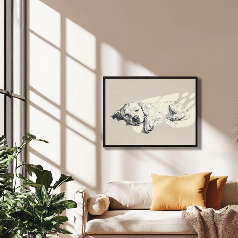 May include: A framed monochrome sketch of a sleeping dog. The artwork is displayed on a cream background, with sunlight casting shadows across the image. The dog is lying down, appearing relaxed. The artwork is hanging above a beige sofa with ochre cushions.