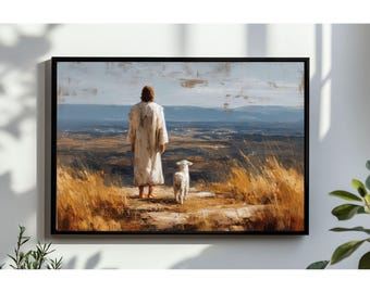 May include: A framed art print depicts a figure in a white robe walking with a lamb. The artwork features a landscape with a blue sky and distant mountains. The foreground shows tall, dry grass.