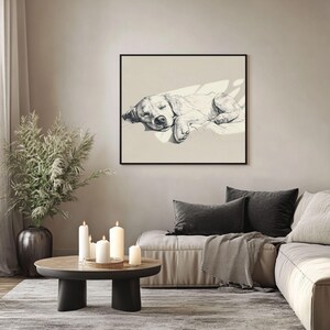 May include: A framed monochrome sketch of a sleeping dog. The artwork hangs on a neutral wall above a sofa with dark grey cushions and a throw. A round coffee table with candles and a decorative plant is in the foreground.