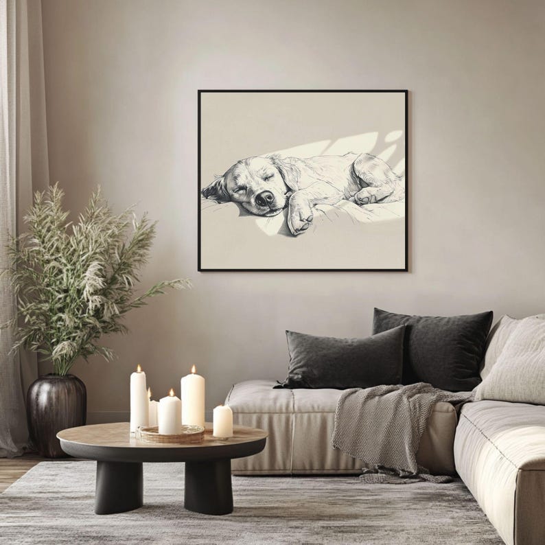 May include: A framed monochrome sketch of a sleeping dog. The artwork hangs on a neutral wall above a sofa with dark grey cushions and a throw. A round coffee table with candles and a decorative plant is in the foreground.