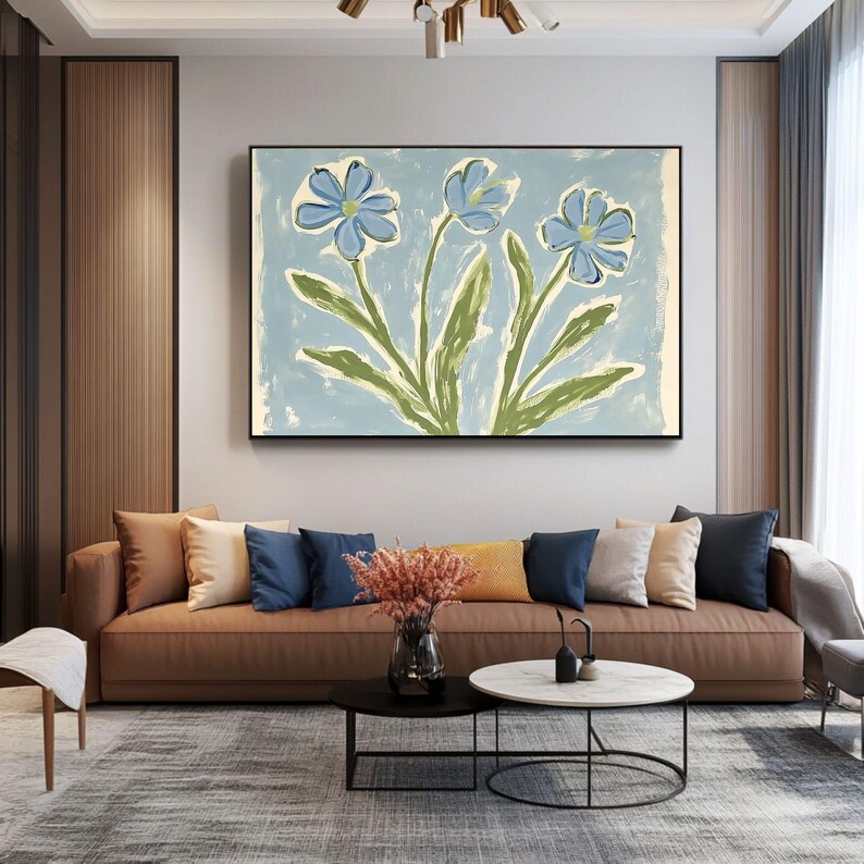 Puede incluir: Una pintura de tres flores azules con tallos y hojas verdes sobre un fondo azul claro. La pintura est&aacute; enmarcada y colgada en una pared sobre un sof&aacute; de cuero marr&oacute;n con almohadas.
