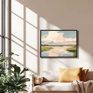 May include: A framed painting depicting a landscape with a river, green grass, and a pink and blue cloudy sky. The painting is in a black frame and is hanging on a white wall.