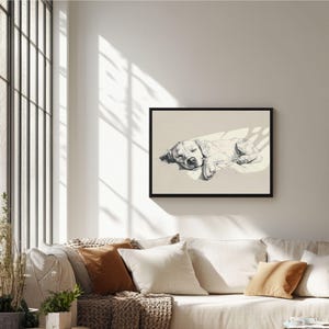 May include: A framed black and white art print depicting a sleeping dog. The dog is shown lying down, with its eyes closed, and is lit by a beam of light. The artwork is displayed above a sofa with several cushions.