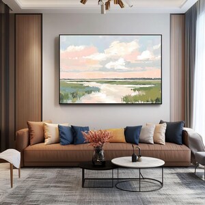 May include: A painting of a landscape with a river flowing through a field of green grass. The sky is a soft pink and blue with white clouds. The painting is framed in a black frame and is hanging on a wall in a living room.