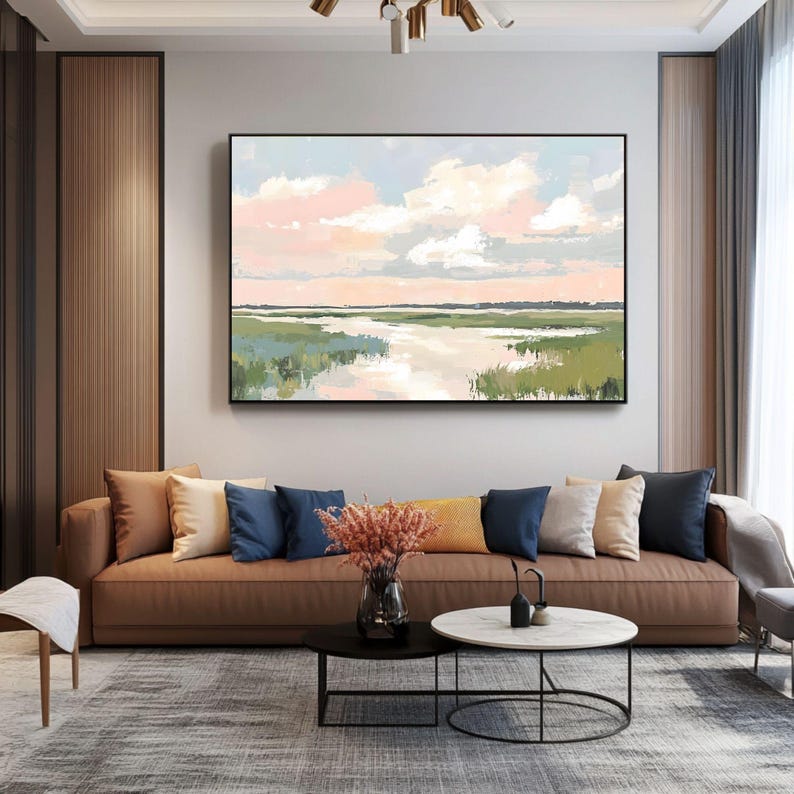 May include: A painting of a landscape with a river flowing through a field of green grass. The sky is a soft pink and blue with white clouds. The painting is framed in a black frame and is hanging on a wall in a living room.