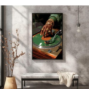 May include: A framed art print depicts a DJ's hand adorned with gold jewelry, manipulating a green vinyl record on a turntable. The artwork features a warm color palette with shades of brown, green, and gold, creating a retro vibe. The image is set against a textured gray wall.