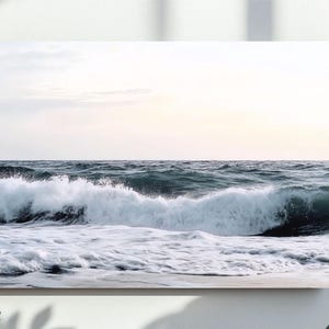 Op de afbeelding: Een foto van een grote golf die breekt op een zandstrand. De golf is wit met een blauwe oceaan erachter. De lucht is lichtblauw met witte wolken.