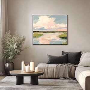 May include: A framed painting depicting a landscape with a lake, green grass, and a pink and blue sky with clouds. The painting is hanging on a wall in a living room featuring a sofa, candles, and a coffee table.