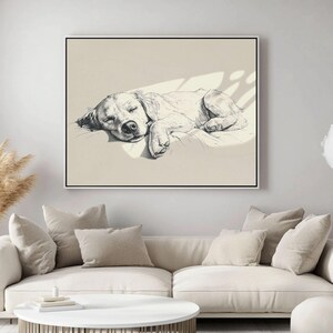 May include: A framed monochrome sketch of a sleeping dog. The dog is depicted lying down, eyes closed, on a light-coloured surface. The artwork is mounted on a wall above a white sofa with beige cushions.