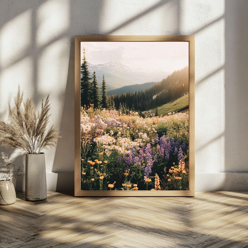 May include: A framed photograph of a mountain meadow with wildflowers in bloom. The meadow is filled with purple, yellow, and white flowers. The mountains in the background are covered in trees and snow. The sun is shining brightly and the sky is a light blue.