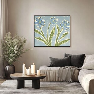 Puede incluir: Una pintura de tres flores azules con tallos y hojas verdes sobre un fondo azul claro. La pintura est&aacute; enmarcada en negro y est&aacute; colgada en una pared de una sala de estar.