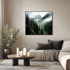 May include: A framed photograph of a misty mountain landscape with a valley of green trees. The photo is hanging on a beige wall above a light brown couch with gray throw pillows and a blanket.