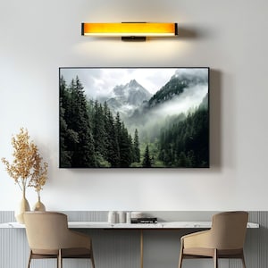 May include: A framed photograph of a misty mountain landscape with lush green trees. The photograph is hanging on a white wall above a wooden table with two chairs.