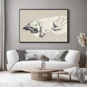 May include: Black and white framed artwork depicting a sleeping dog. The dog is resting on a pillow. The artwork is displayed above a white sofa with grey cushions and a wooden coffee table.