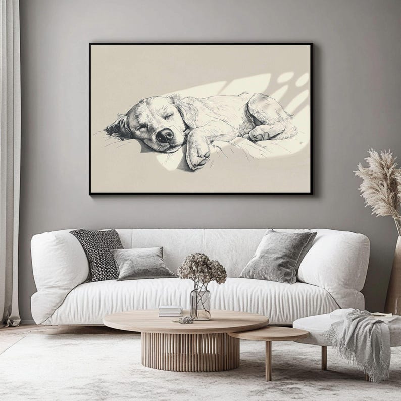 May include: Black and white framed artwork depicting a sleeping dog. The dog is resting on a pillow. The artwork is displayed above a white sofa with grey cushions and a wooden coffee table.