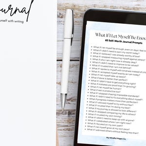 May include: A white journal with the word "Journal" in cursive, a white pen, and a smartphone displaying "What If I Let Myself Be Enough" journal prompts. The journal and phone are on a light wood surface.