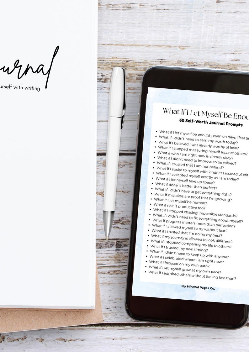 May include: A white journal with the word "Journal" in cursive, a white pen, and a smartphone displaying "What If I Let Myself Be Enough" journal prompts. The journal and phone are on a light wood surface.