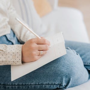 May include: A person writing in a white notebook with a pencil. The person is wearing a cream-colored shirt with black polka dots and blue jeans. The person is sitting on a white surface.