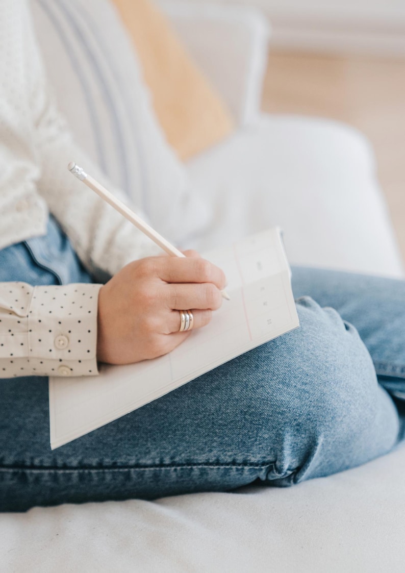 May include: A person writing in a white notebook with a pencil. The person is wearing a cream-colored shirt with black polka dots and blue jeans. The person is sitting on a white surface.