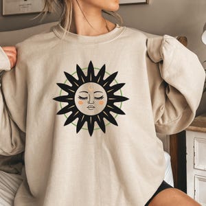 Puede incluir: Una sudadera beige con un gráfico de sol negro y verde. El sol tiene una cara con los ojos cerrados y una luna creciente por encima.