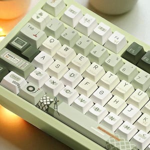 May include: A light green mechanical keyboard with white and green keycaps. The keyboard has a variety of symbols and text on the keys, including numbers, letters, and special characters. The keyboard is angled towards the viewer.
