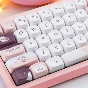 May include: A pink mechanical keyboard with white and pink keycaps. The keycaps have white lettering and some feature custom designs. The keyboard has a pink frame and the text "Peach Choc" is printed on the spacebar.