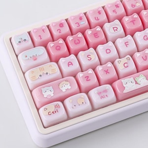 May include: A close-up of a keyboard with pink and white cat-themed keycaps. The keycaps feature cat faces and various symbols and letters. The keyboard has a white frame and a light pink base.