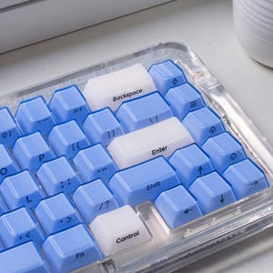 May include: A compact, clear keyboard with light blue and white keycaps. The keys are arranged in a grid, with labels like "Backspace", "Enter", and "Shift". The keyboard sits on a white surface, with a small potted plant in the background.