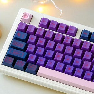 May include: A white mechanical keyboard with purple, blue, and pink keycaps. The keycaps have red lettering. The keyboard is on a white surface with a small succulent plant in a white pot and string lights in the background.