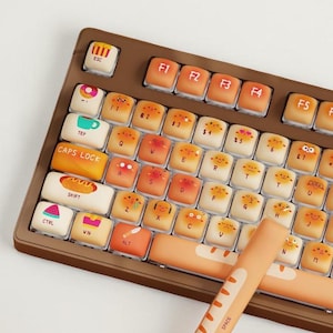May include: A brown mechanical keyboard with keycaps designed to look like bread and pastries. The space bar is a long, orange baguette. The keys are a mix of orange, white, and brown, with various food-related icons.