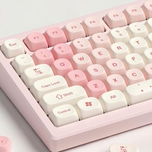 May include: A pastel pink mechanical keyboard with white and pink keycaps. The keycaps are arranged in a standard layout, with the letters and symbols printed in a clean, modern font. Several keycaps are detached and lying next to the keyboard.
