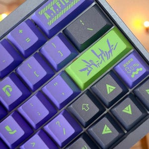 May include: A close-up of a mechanical keyboard with purple and gray keys. The keyboard has a silver frame and features custom keycaps with Japanese text and the text "A.T. FIELD". The keycaps are purple, green, and gray.