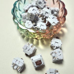 May include: A collection of white mechanical keyboard switches with brown stems and gold pins, scattered in and around a decorative, multi-colored glass bowl. The switches are small, square, and have a light gray housing.