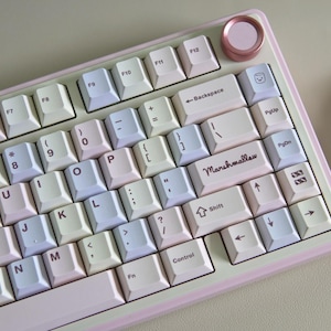May include: A mechanical keyboard with a pastel color scheme featuring white, light pink, and light blue keycaps. The enter key is labeled with the word Marshmallow in a cursive font. A circular metallic pink knob is located in the top right corner.