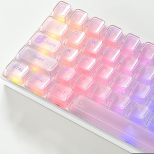 May include: A mechanical keyboard featuring translucent, iridescent keycaps with a glossy finish. The keys display standard QWERTY lettering in a purple font, illuminated by a soft, multi-colored internal glow that shifts between pink, orange, and blue hues.